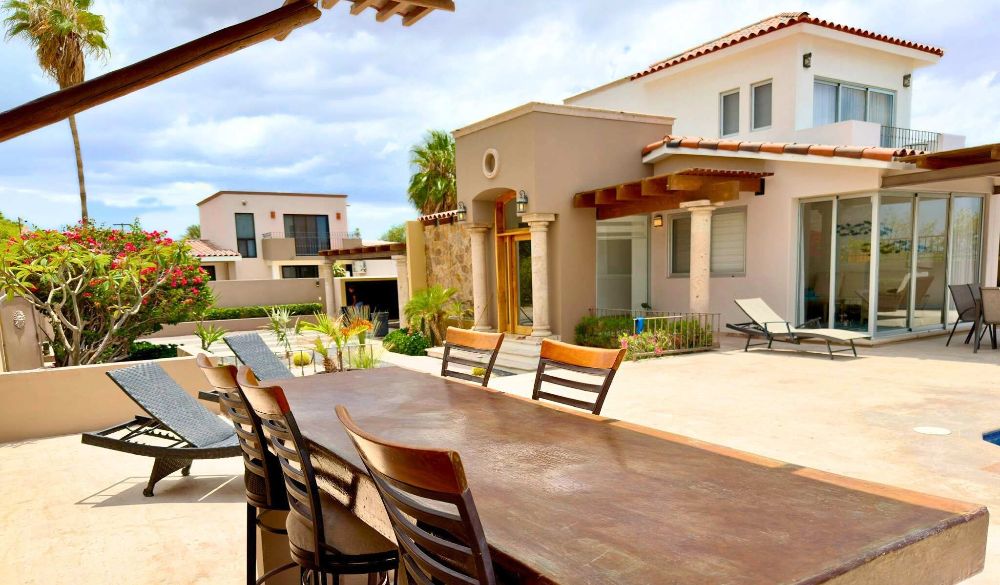 A modern villa with a patio area featuring a dining table and chairs, a covered seating area, and a poolside lounge.