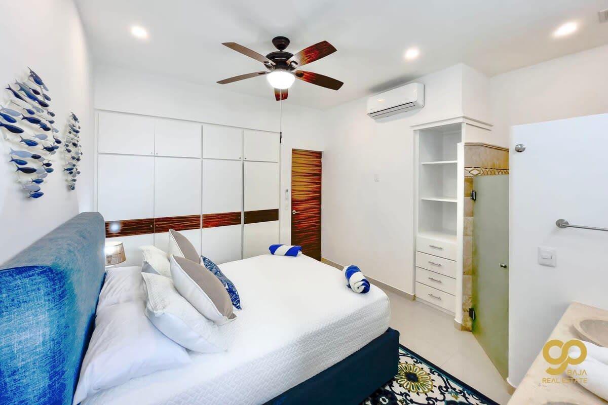 A modern hotel room with a bed, a ceiling fan, a wardrobe, and a bathroom with a sink and mirror.