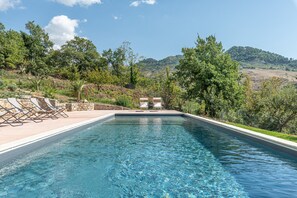 A rectangular swimming pool with clear blue water, surrounded by a stone deck and white lounge chairs.