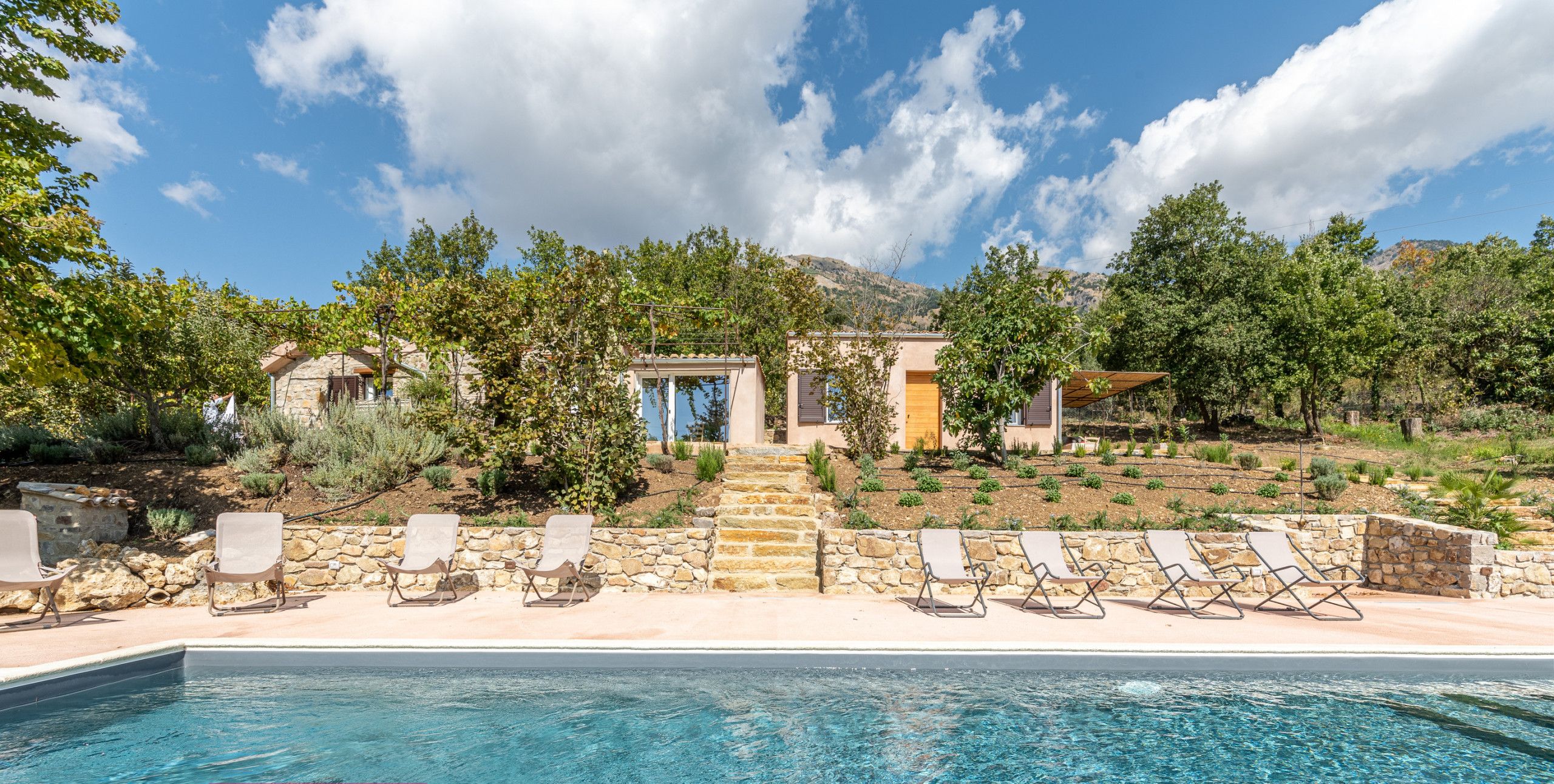 A poolside view of a property with a stone pathway leading to a building, surrounded by trees and a clear sky.