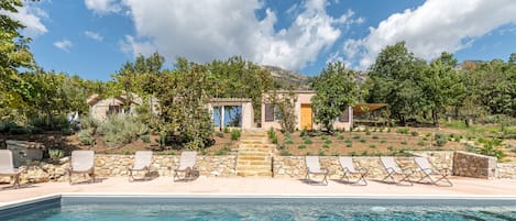 A poolside view of a property with a stone pathway leading to a building, surrounded by trees and a clear sky.