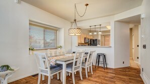 Een eetgedeelte met een houten tafel en witte stoelen, een keuken op de achtergrond en een hanglamp.