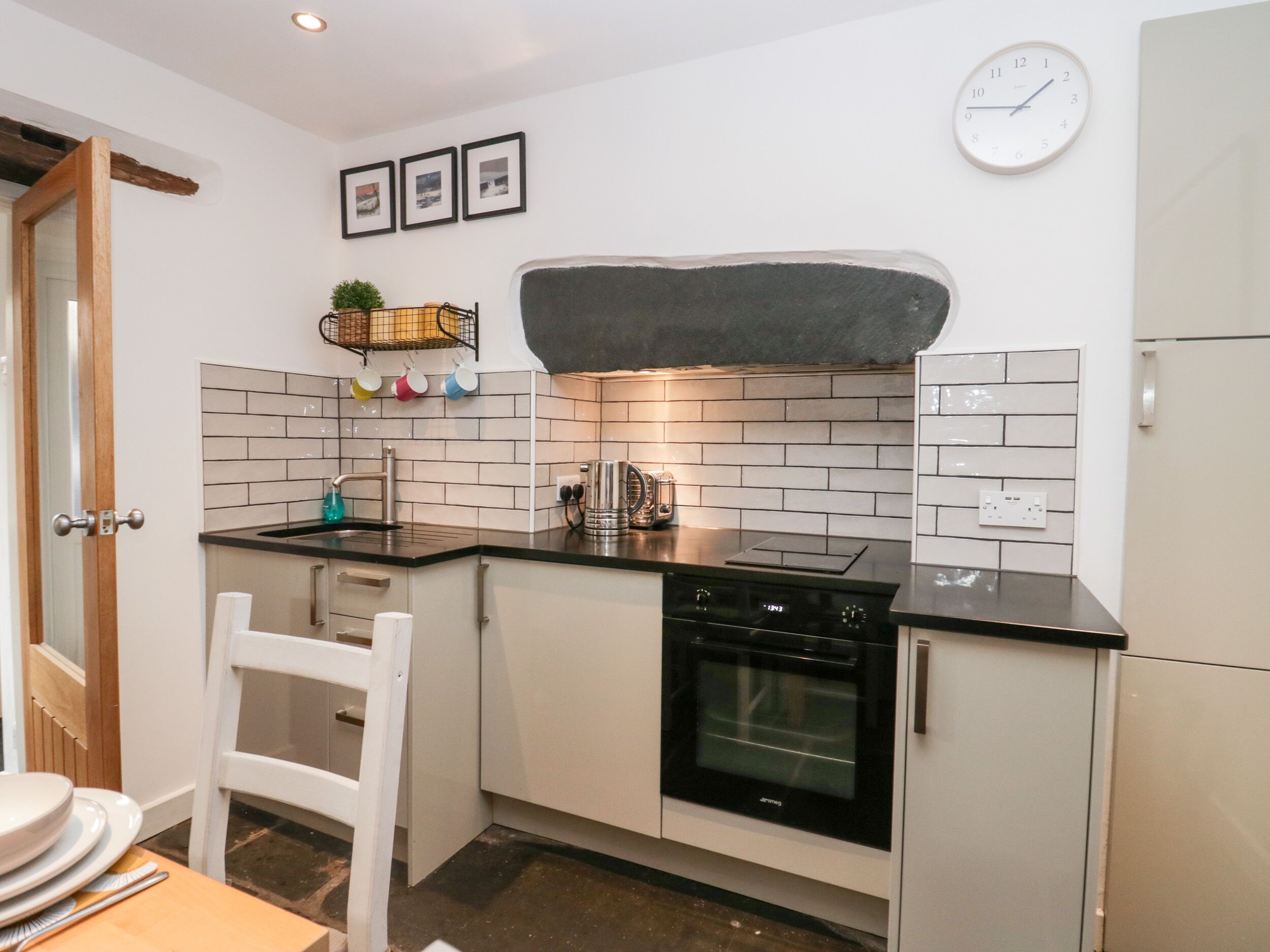 Een kleine keuken met witte kastjes, een zwart aanrecht, een inbouwoven en een hangend horloge.