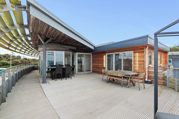 A covered outdoor area with a wooden deck, a seating area with tables and chairs, and a glass door leading to a building.