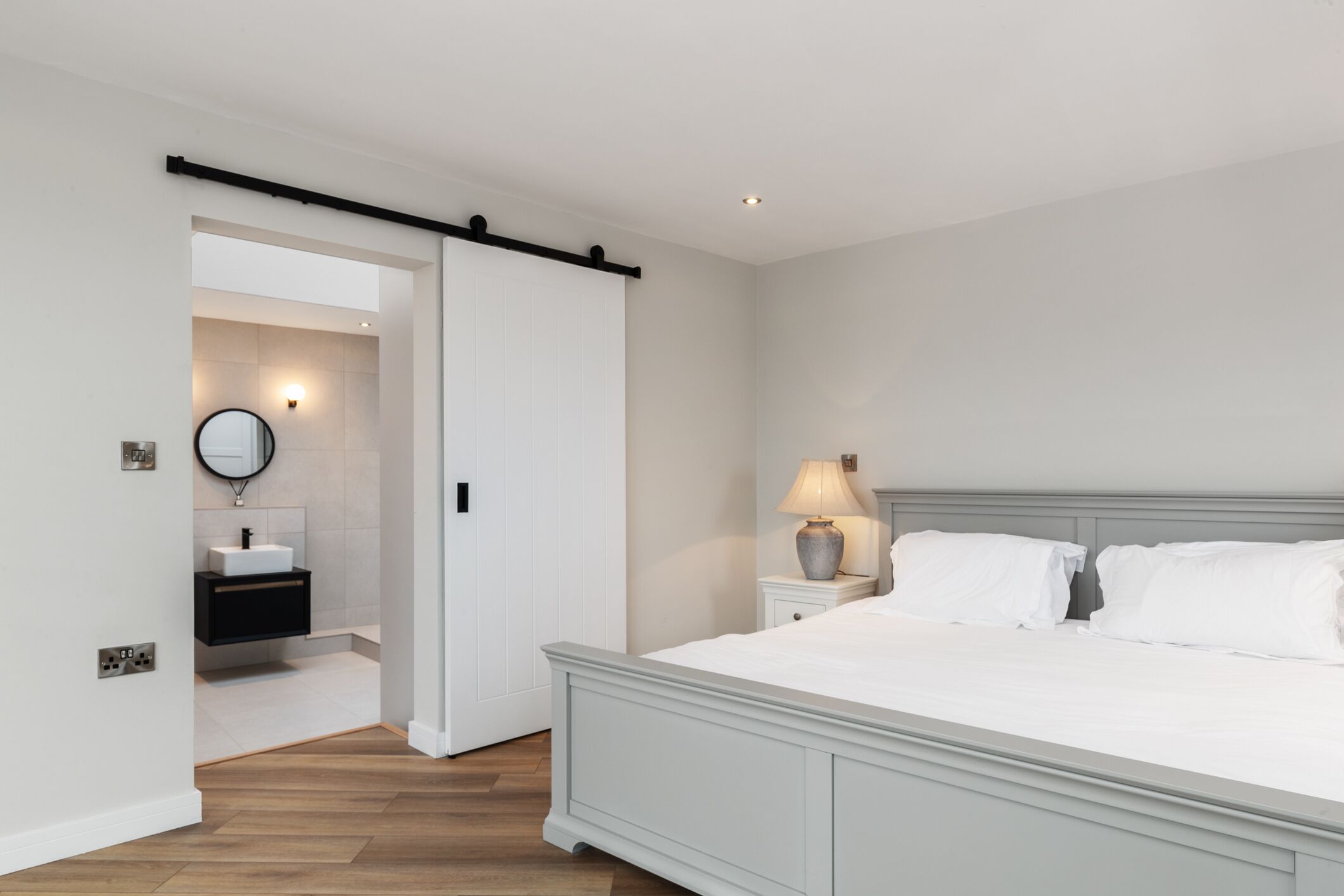 Un dormitorio con una cama grande, una mesita de noche con lámpara, una mesita auxiliar y una puerta corredera que da al baño, además de un espejo montado en la pared.