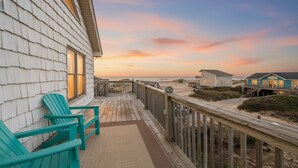 A wooden deck with two green chairs, a wooden railing, and a view of the beach and houses in the background.