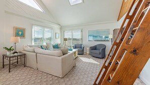 A spacious living room with a large sofa, a coffee table, and two armchairs. There are large windows with blinds, a painting on the wall, and a wooden ladder leaning against the wall.