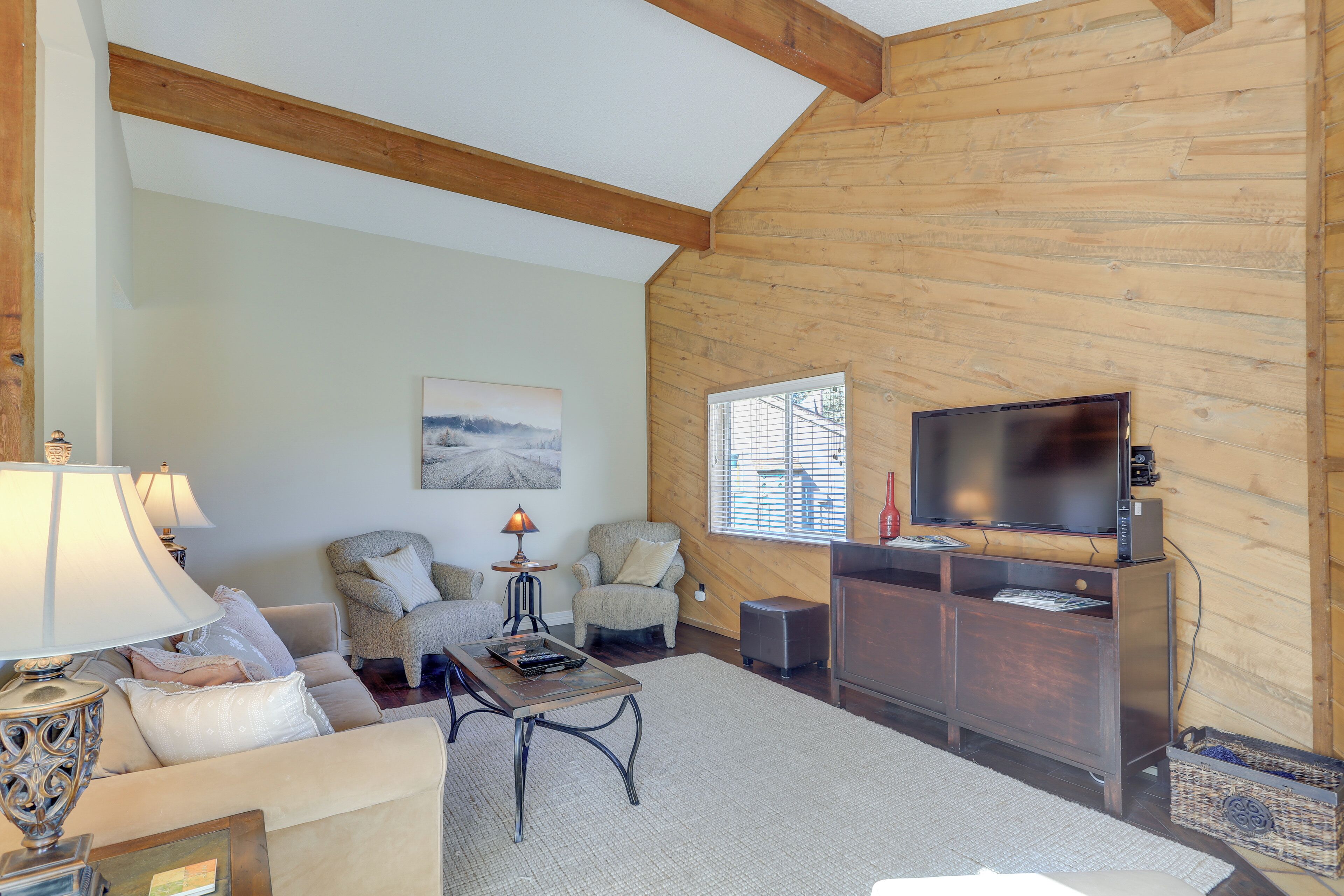 A cozy living room with a wooden ceiling, a flat-screen TV on a wooden stand, a sofa, two armchairs, a coffee table, and a lamp.