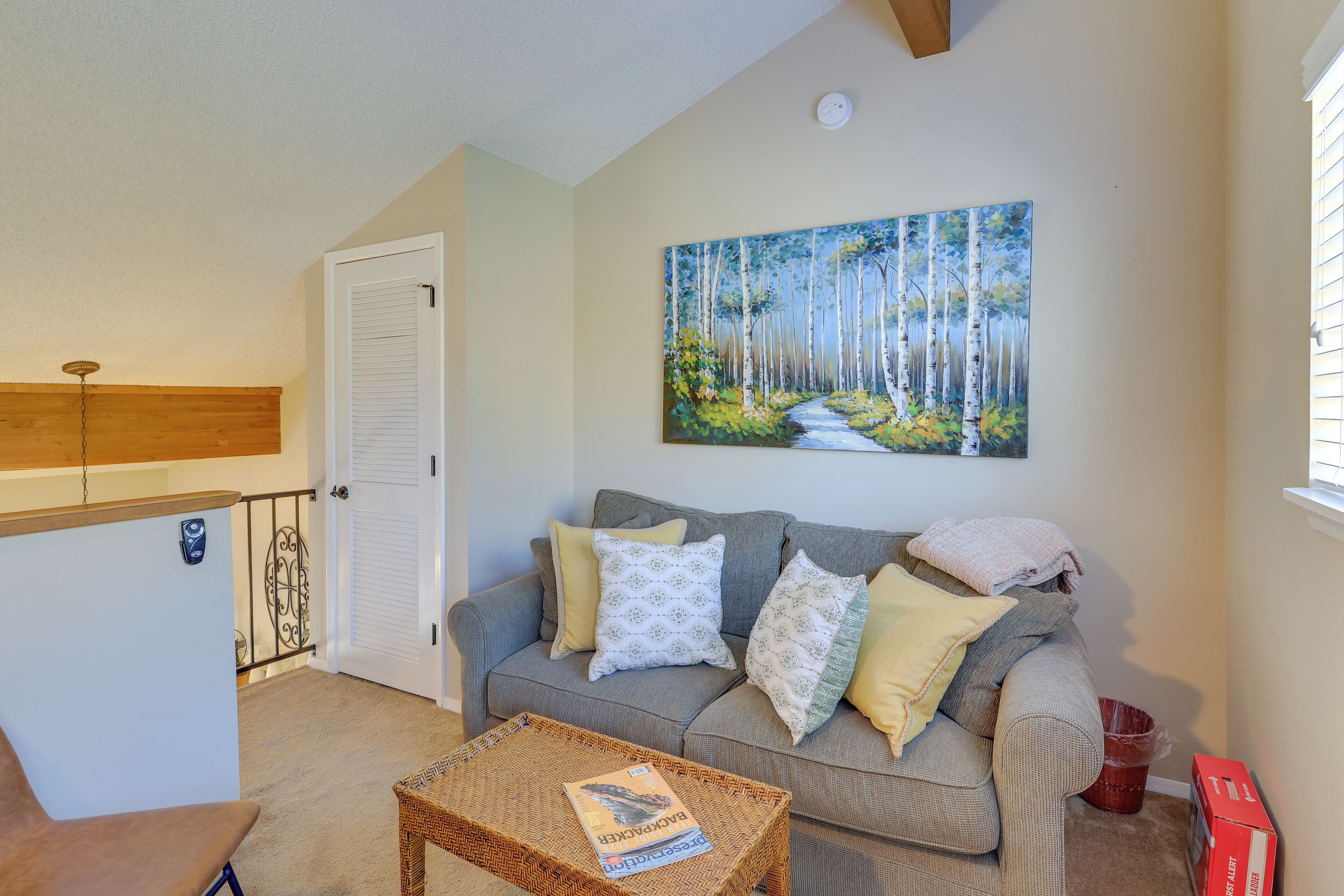A living room with a grey sofa, a painting of a forest, a wicker coffee table, and a door with a white shutter.