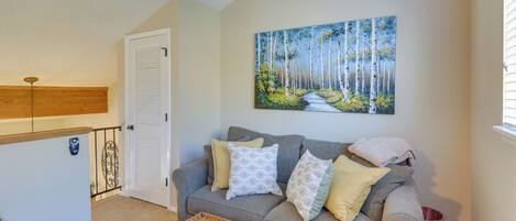 A living room with a grey sofa, a painting of a forest, a wicker coffee table, and a door with a white shutter.