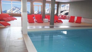 Piscina interior con sillas reclinables rojas y vistas a montañas nevadas a través de amplias ventanas.