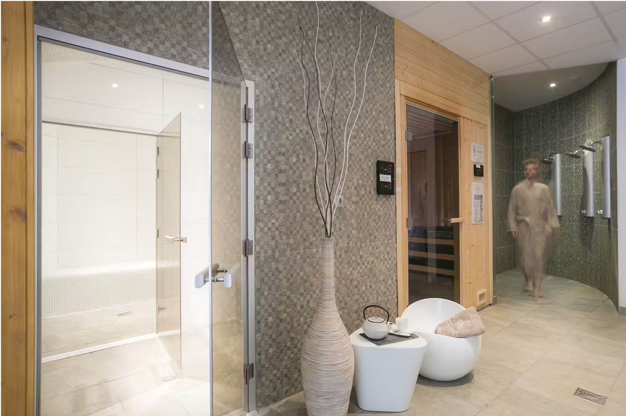 A modern bathroom with a glass shower enclosure, a textured wall, a tall vase, and a seating area with a teapot.