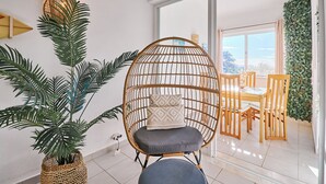A room with a wicker chair, a dining table with chairs, a potted plant, and a view of the outdoors through a large window. - Apartment Buenos Momentos (Carvoeiro)