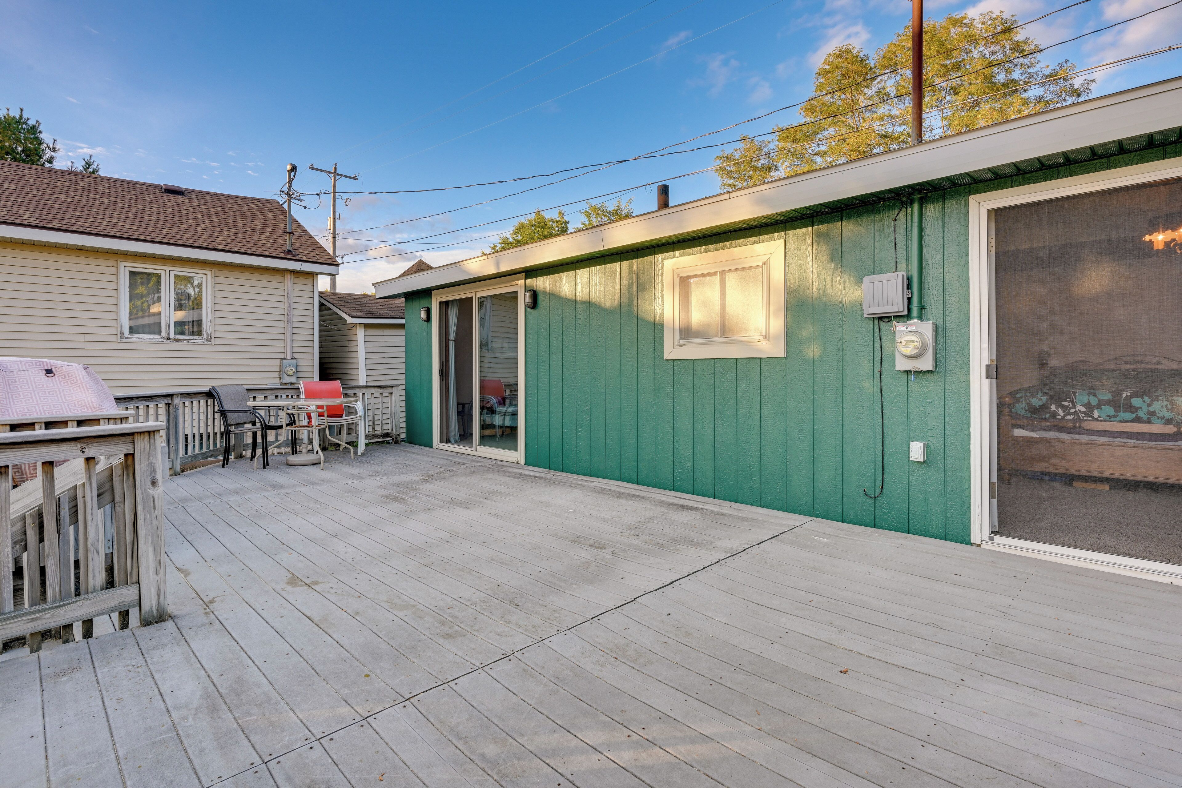 A wooden deck with a grill and chairs, a green building with a garage door, and a clear sky.