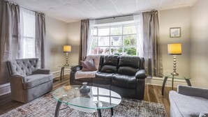 A living room with a grey armchair, a black leather sofa, a glass coffee table, and two lamps on side tables. - Central Auburn Apartment w/ Porch: Pets Welcome! (Auburn)