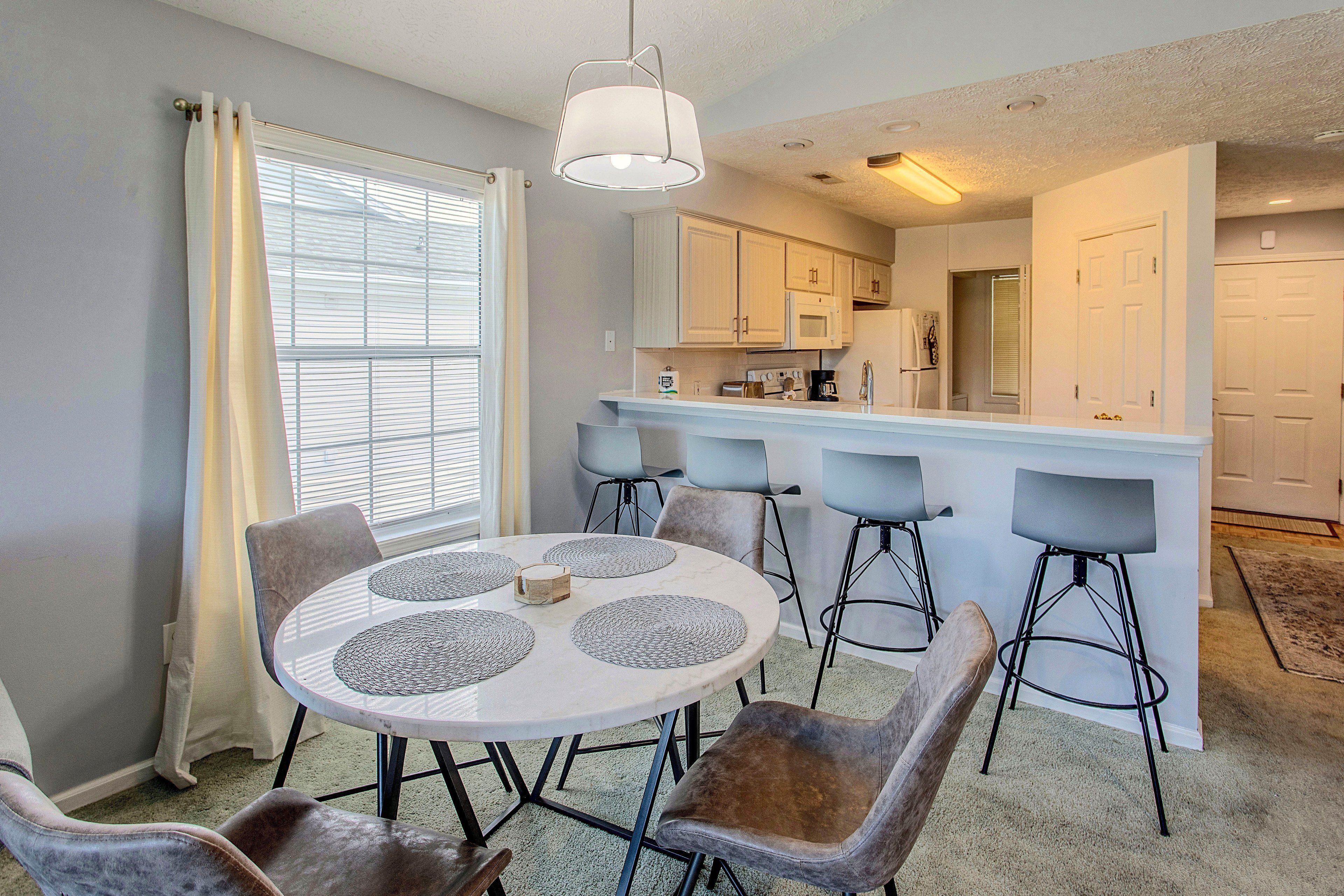 A dining area with a round table, four chairs, and a kitchen in the background.