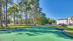 A green outdoor area with a curved path, surrounded by tall trees and a building in the background. - Myrtle Beach Oasis Condo w/ Pool Access! (Myrtle Beach)
