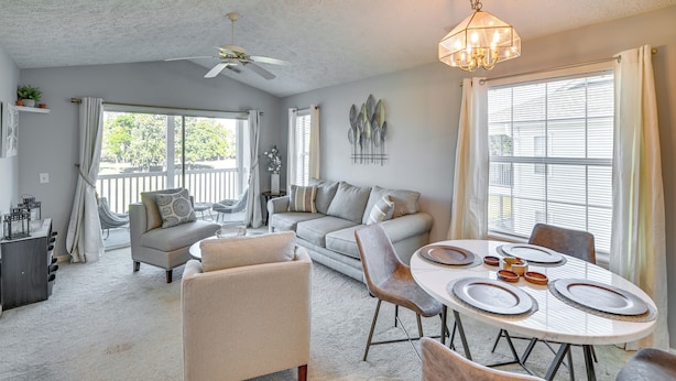 A spacious living room with a sofa, dining table, and chairs. There is a ceiling fan, a chandelier, and large windows with curtains. - Myrtle Beach Oasis Condo w/ Pool Access! (Myrtle Beach)