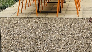 Une table en bois, entourée de chaises orange, est placée sur une terrasse dont le sol est recouvert de gravier.