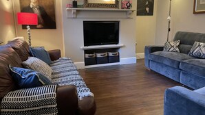 Un salon avec un canapé en cuir marron, un canapé bleu, un téléviseur à écran plat, une cheminée et un lampadaire rouge.