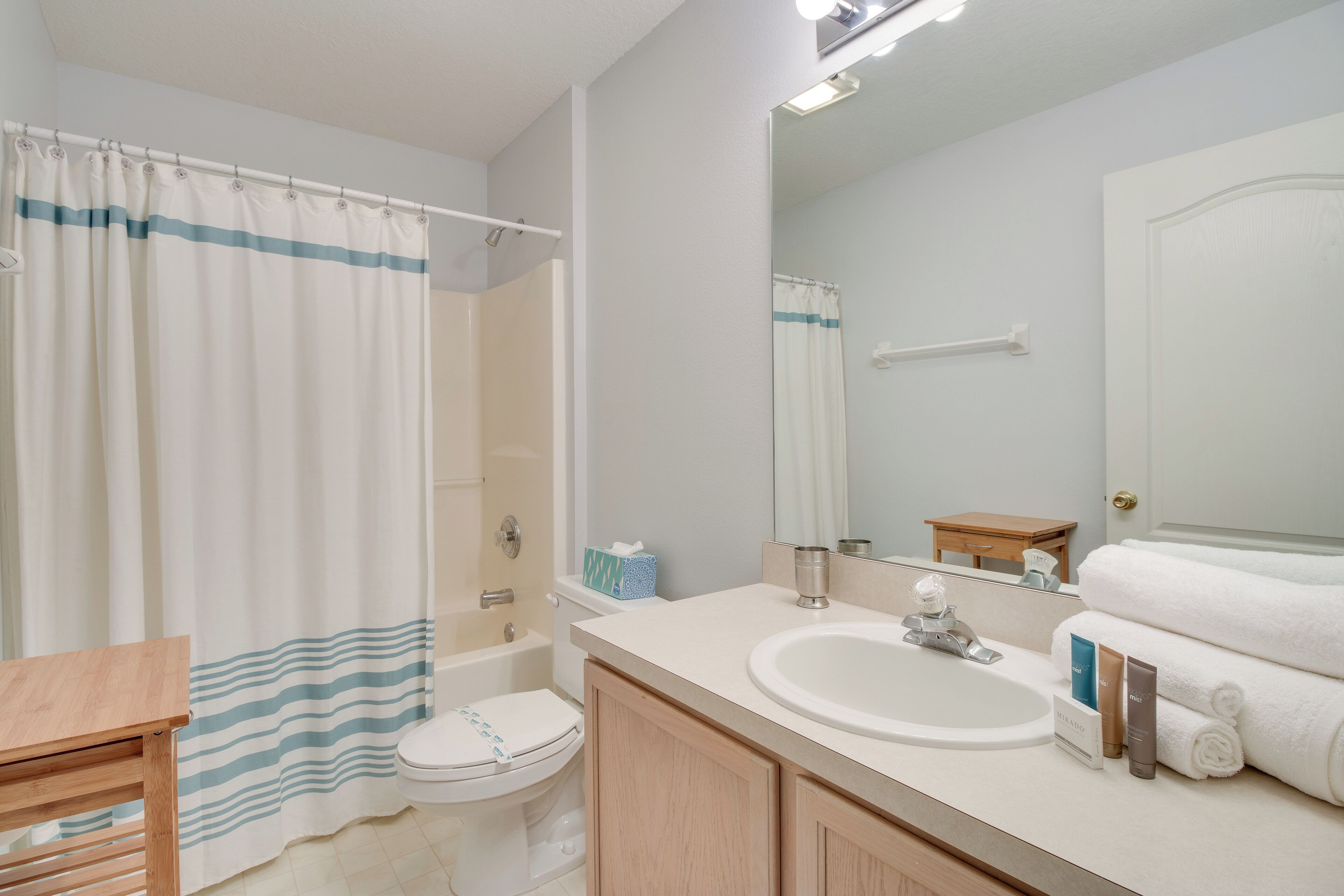 A bathroom with a shower curtain, toilet, sink, mirror, and a small wooden table.