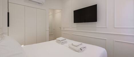 A hotel room with a bed, a flat-screen TV mounted on the wall, and an air conditioning unit above the bed.