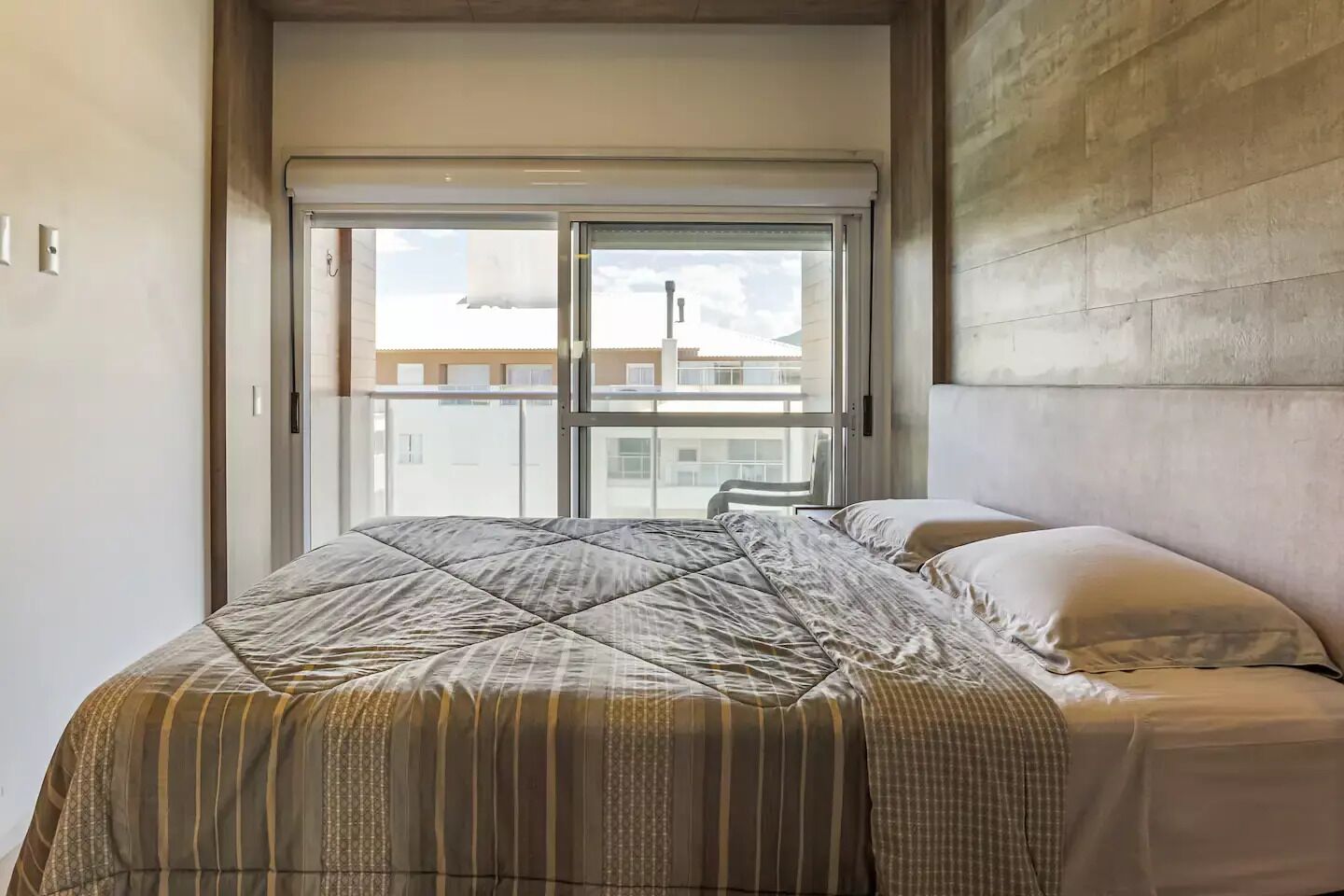 Un dormitorio con un ventanal grande, una cama con ropa de cama a rayas y un cabecero sencillo.