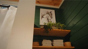 A bathroom with a green wall, a towel rack, a framed picture of a horse, and a shelf with folded towels and a plant. - White Oak Creek Cabin (Harrison)