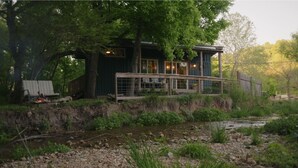 A rustic cabin with a wooden deck, a small fire, and a river in front. - White Oak Creek Cabin (Harrison)