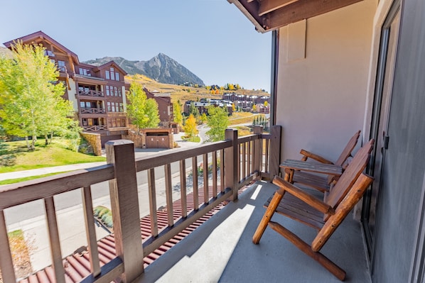 A balcony with wooden railings and chairs, overlooking a town with buildings and a mountain in the background.