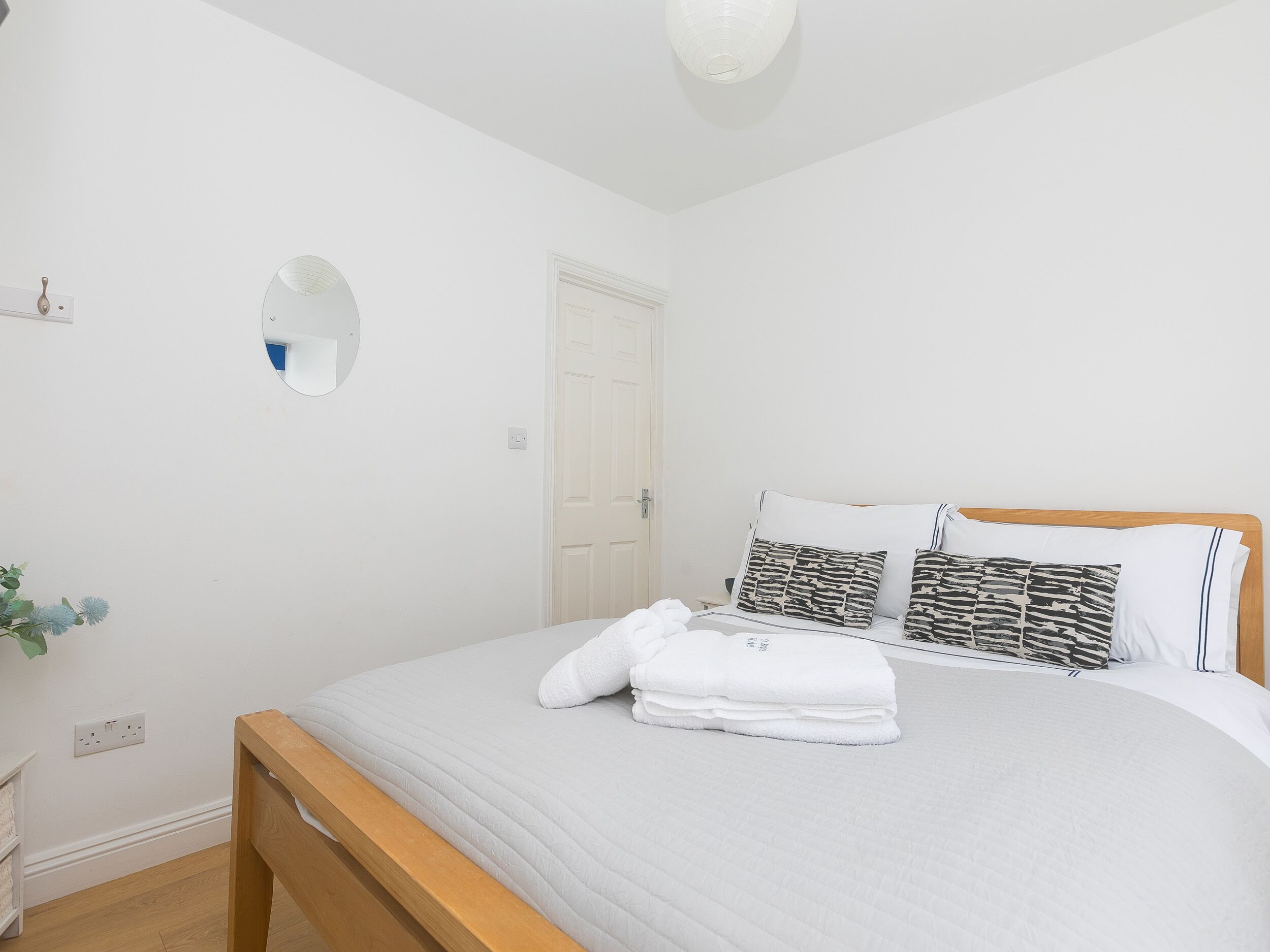 A bedroom with a bed, white walls, a wooden headboard, a round mirror, and a white door.