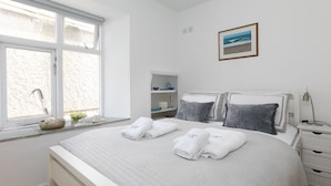A bedroom with a bed, a bedside table, a lamp, a shelf with toiletries, and a window with a view of the sea. - Harbour Side (St Ives)