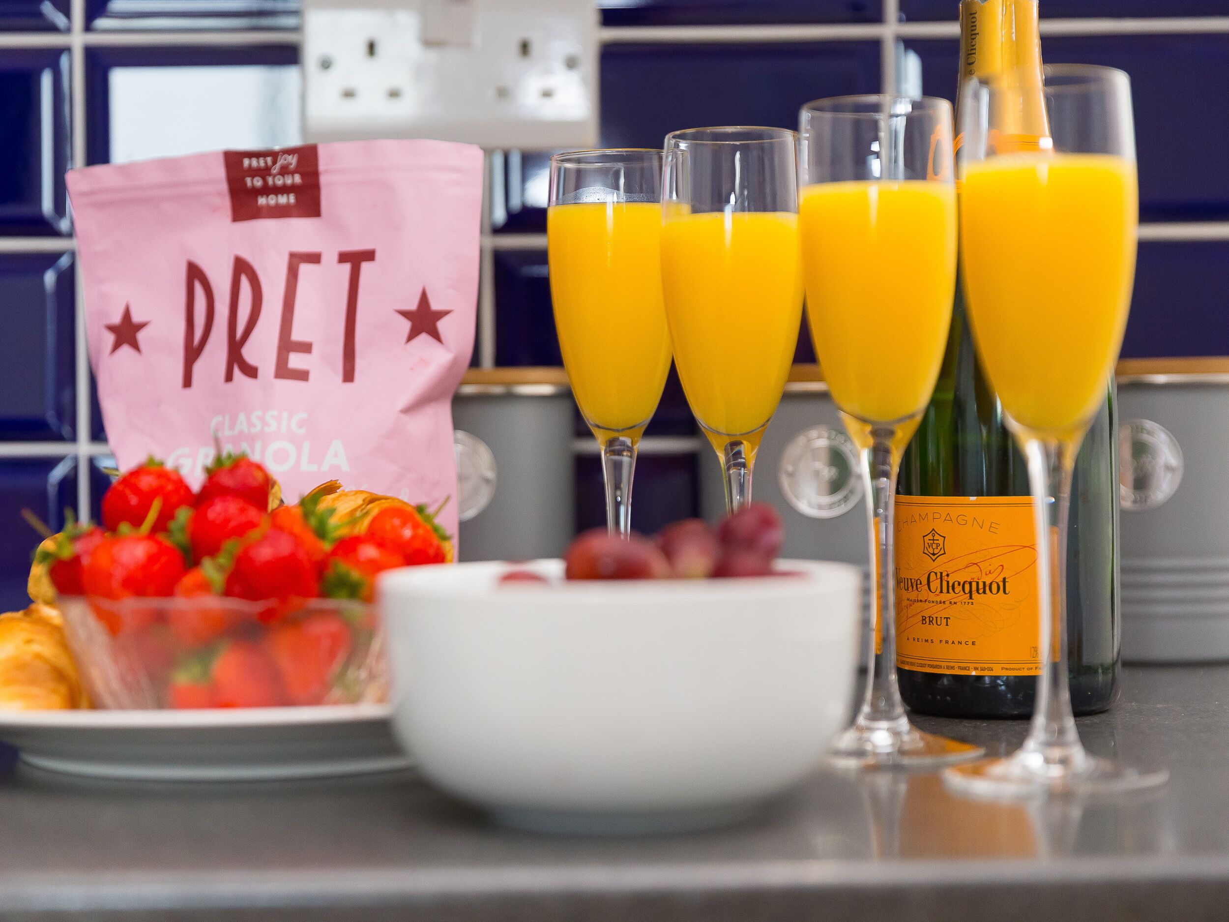 Un comptoir de cuisine avec un sac étiqueté « DRET », trois verres de jus d’orange, une bouteille de champagne et un bol de fraises.
