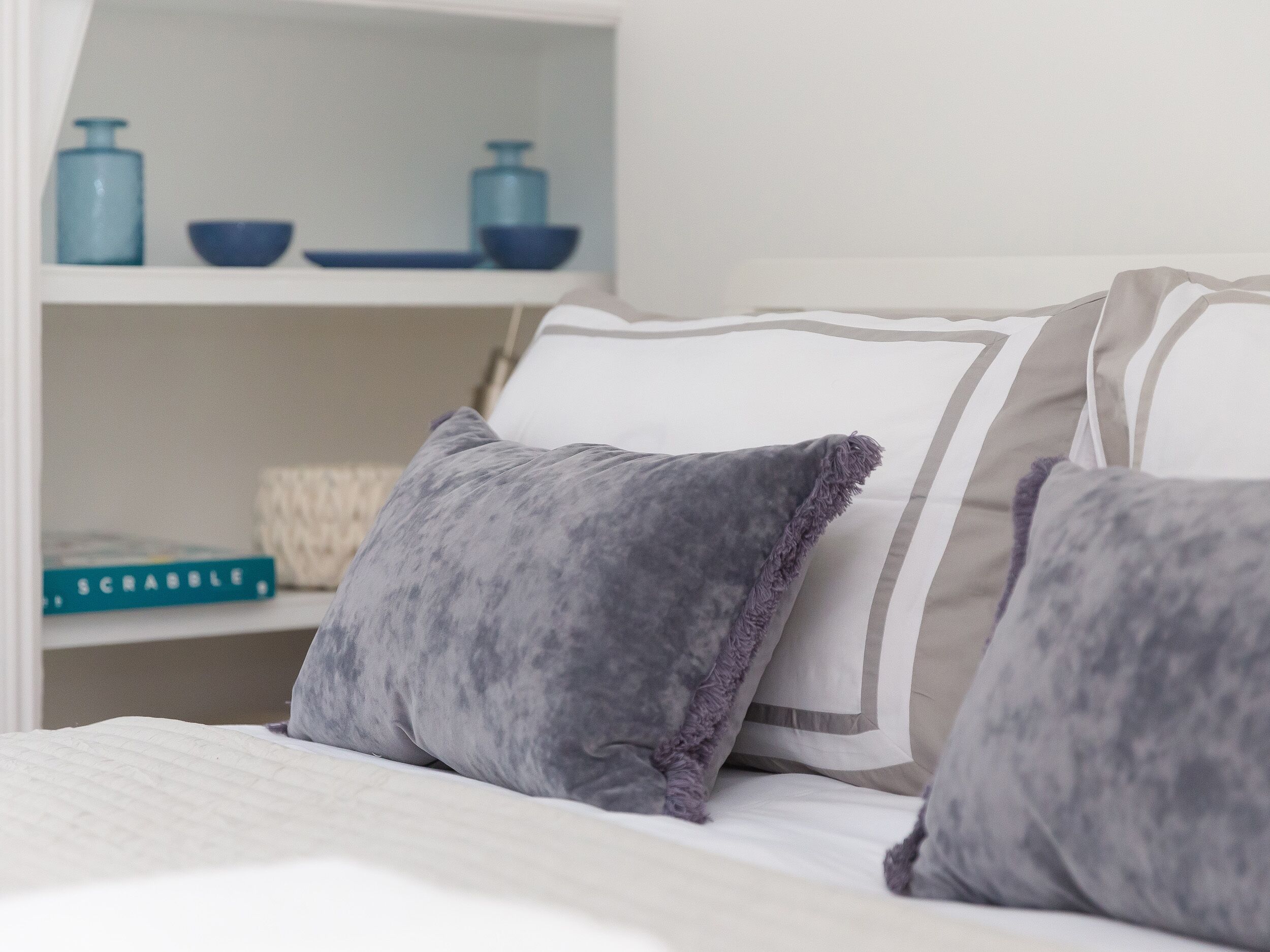 A neatly made bed with grey and white pillows, a white headboard, and a shelf with decorative items and a book.