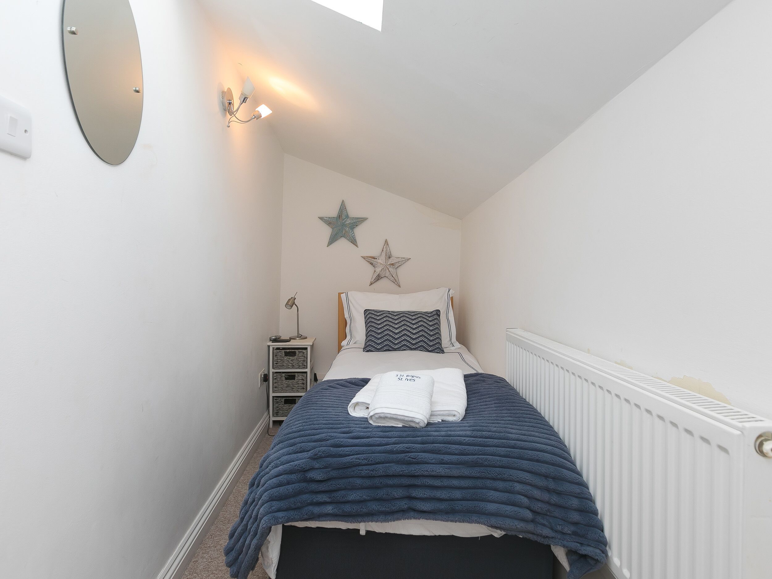 A narrow, white-walled bedroom with a bed, a star decoration, a nightstand, and a wall-mounted light fixture.