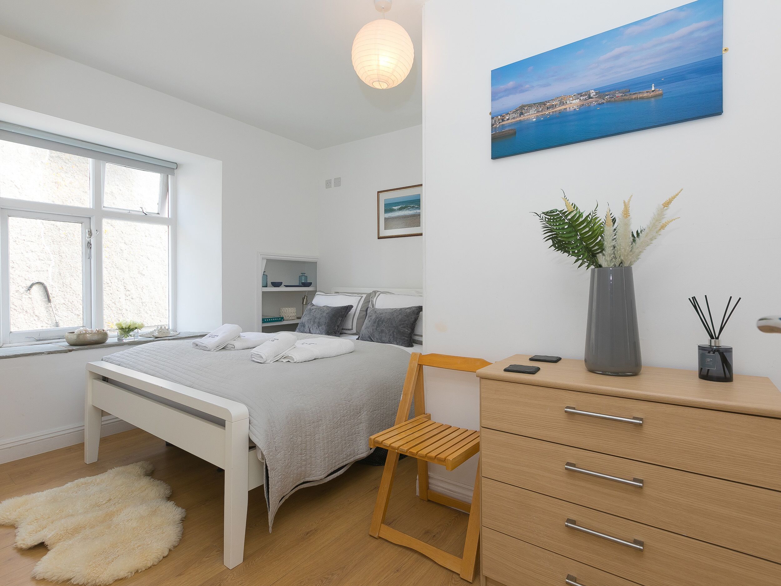 A modern bedroom with a bed, bedside table, dresser, and a painting of a coastal scene.