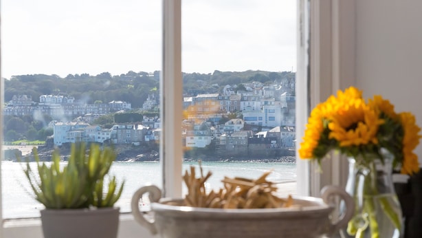 A view of a coastal town through a window, with a bowl of dried seaweed and a vase of yellow flowers on a windowsill. - Harbour Side (St Ives)