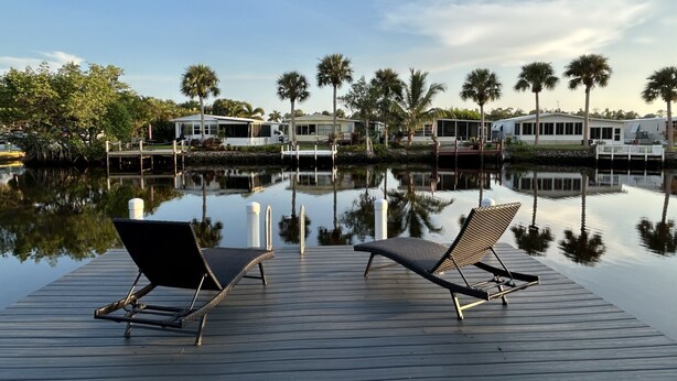 A wooden dock with two lounge chairs facing a calm water body, surrounded by palm trees and a row of mobile homes.