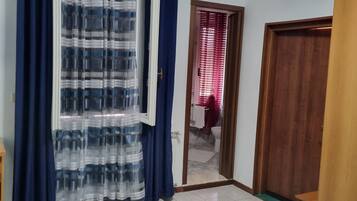 A room with a large window covered by blue and white curtains, a wooden door, a patterned rug, and a bed with a floral bedspread.
