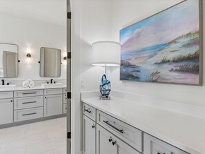A modern bathroom with a white vanity, two mirrors, a painting of a landscape, and a blue decorative object on the counter.
