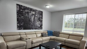 Uma sala de estar com um sofá seccional bege, uma mesa de centro preta e uma grande pintura em preto e branco de um horizonte urbano na parede.