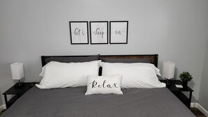 Cama bem arrumada com roupa de cama branca e cinza, cabeceira de madeira e mesa de cabeceira com uma luminária e uma planta pequena.