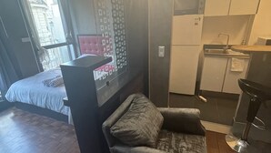 A compact living space with a bed, a small sofa, a kitchenette, and a window with a view of buildings.