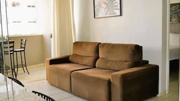 A brown sofa in a room with a tiled floor, a dining table with chairs, and a framed picture on the wall.