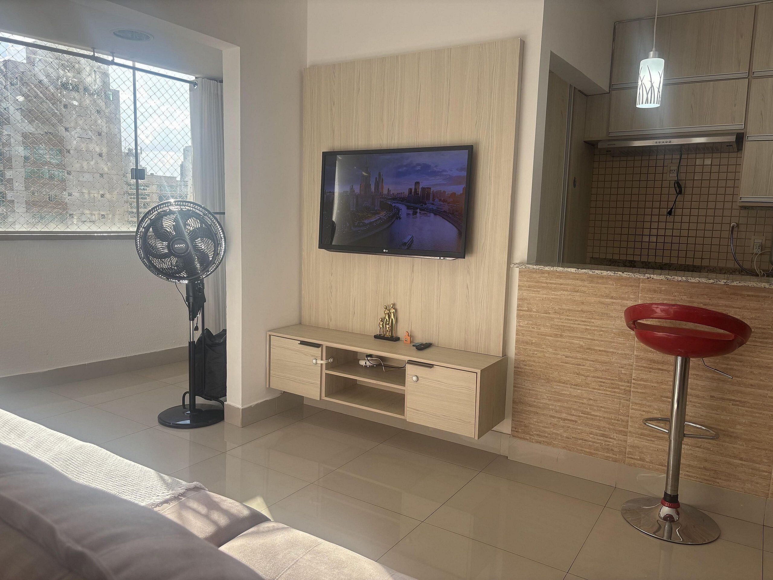Una sala de estar moderna con un televisor de pantalla plana montado en una pared de madera, un ventilador de pie y un taburete rojo.