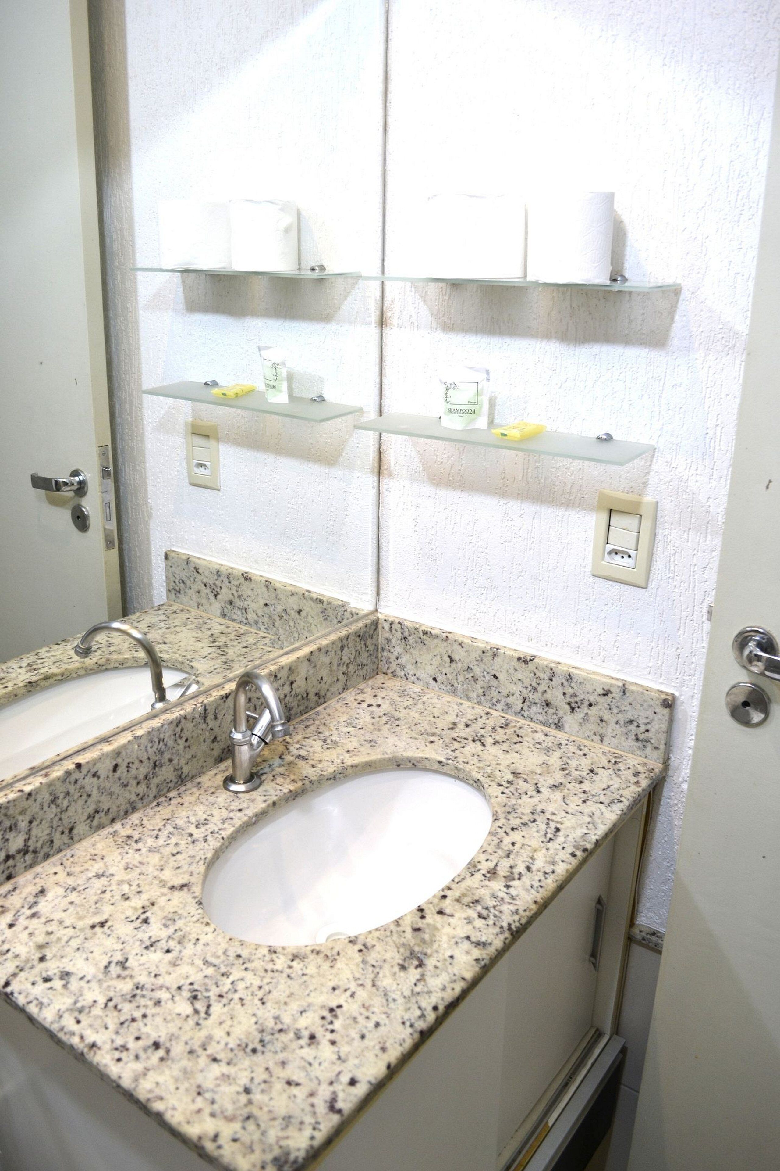 A bathroom with a granite countertop, two sinks, a mirror, and glass shelves above the sinks.