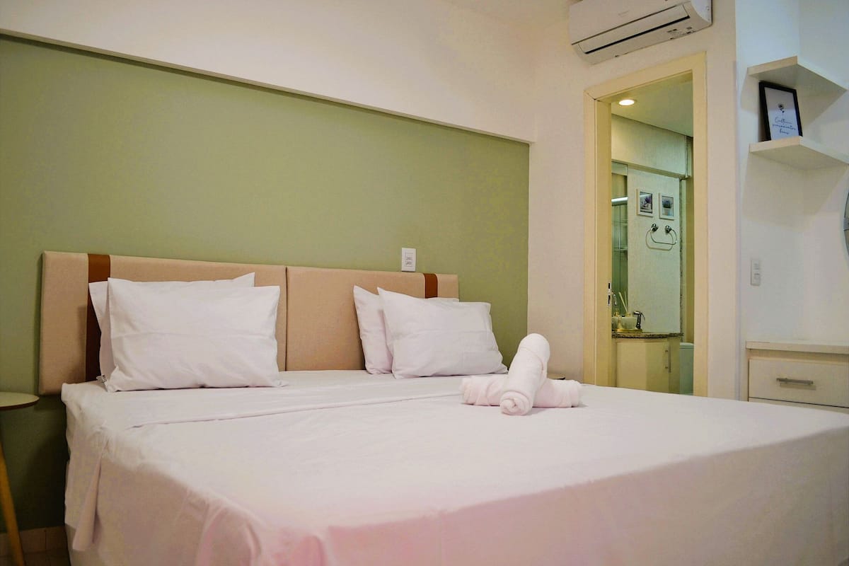 Una cama bien hecha con sábanas y almohadas blancas, una mesita de noche y un aire acondicionado de pared.