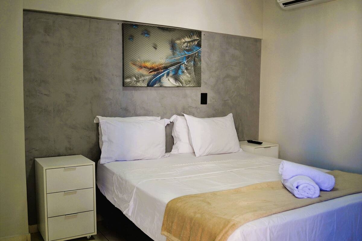 Une chambre d’hôtel avec un lit, du linge de lit blanc, une tête de lit ornée d’une œuvre d’art en plumes, une table de chevet et une serviette roulée.