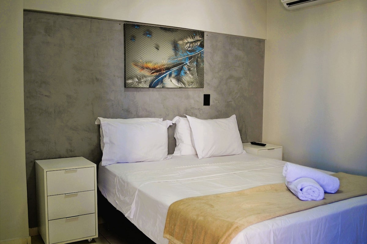 Habitación de hotel con cama, ropa blanca, cabecero con una obra de arte de plumas, mesita de noche y una toalla enrollada.