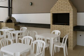 An outdoor patio with a brick oven and grill, white plastic chairs, and tables. - UNIQUE RESIDENCE (Goiania)